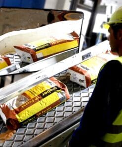 Pracownik w żółtej kamizelce odblaskowej przy taśmie produkcyjnej, z workami materiałów budowlanych marki Weber TP Farvet. Zwraca uwagę na odbicie w lustrze, co podkreśla znaczenie bezpieczeństwa pracy w środowisku przemysłowym.