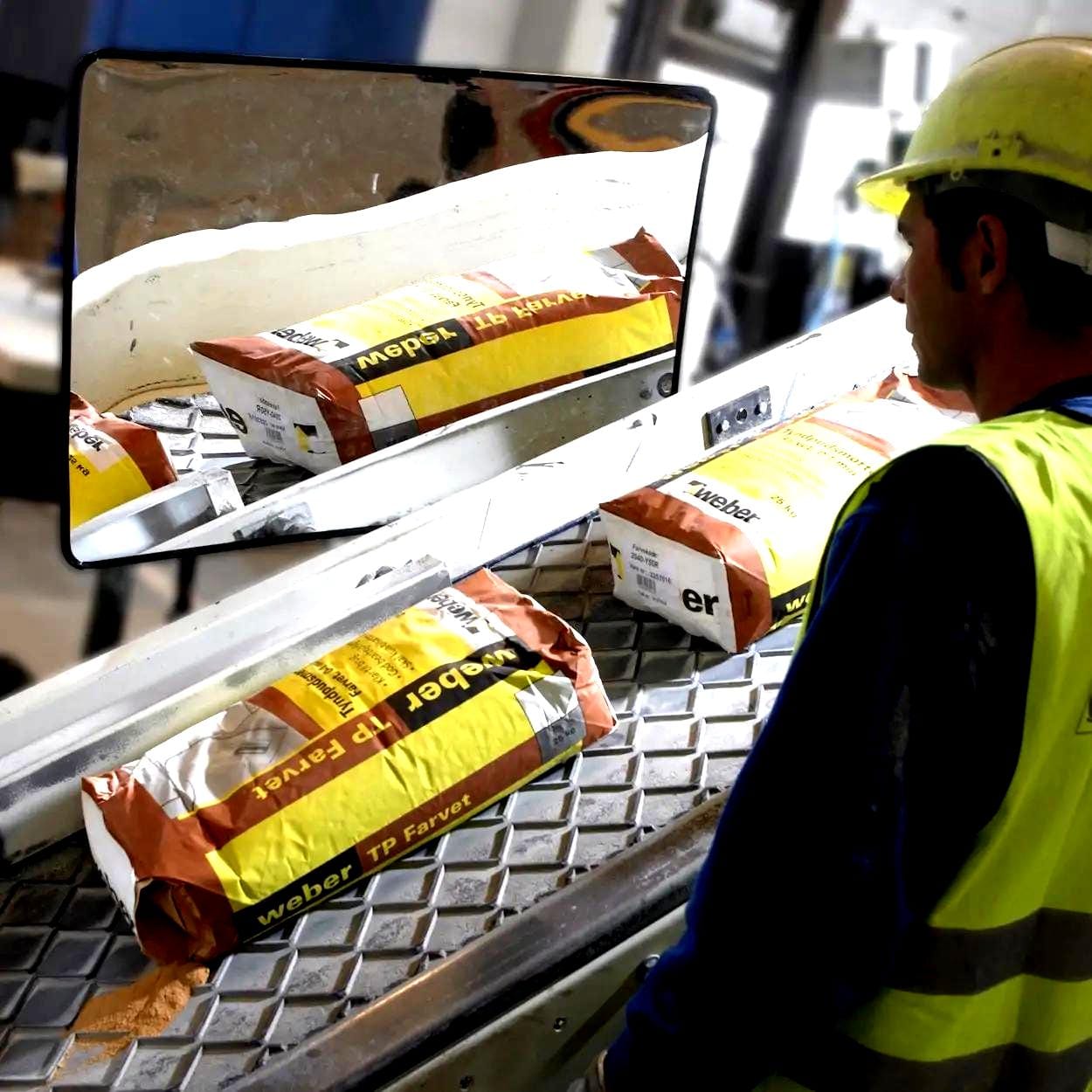 Pracownik w żółtej kamizelce odblaskowej przy taśmie produkcyjnej, z workami materiałów budowlanych marki Weber TP Farvet. Zwraca uwagę na odbicie w lustrze, co podkreśla znaczenie bezpieczeństwa pracy w środowisku przemysłowym.