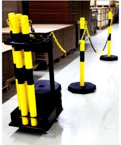 Zestaw znaków ostrzegawczych w magazynie, składający się z żółtych słupków z czarnymi pasami, zamocowanych na octagonowych podstawach. Słupki są połączone czarnym łańcuchem, stojąc na gładkiej podłodze. W tle widoczne kartony, co sugeruje przemysłowe zastosowanie i znaczenie dla bezpieczeństwa pracy.