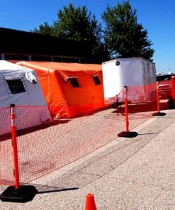 Obszar zabezpieczony siatką w pobliżu białych i pomarańczowych namiotów, w tym białej przyczepy, otoczony konusami. Ujęcie na parkingu pod jasnym niebem. Elementy wskazujące na środki awaryjne i bezpieczeństwo pracy w terenie.