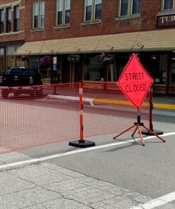 Ulica zamknięta dla ruchu, zabezpieczona pomarańczowymi barierkami i znakiem ostrzegawczym. W tle widoczny budynek z witrynami i jazda dostawcza. W kontekście bezpieczeństwa pracy, oznaczenia te informują o tymczasowych ograniczeniach w ruchu.