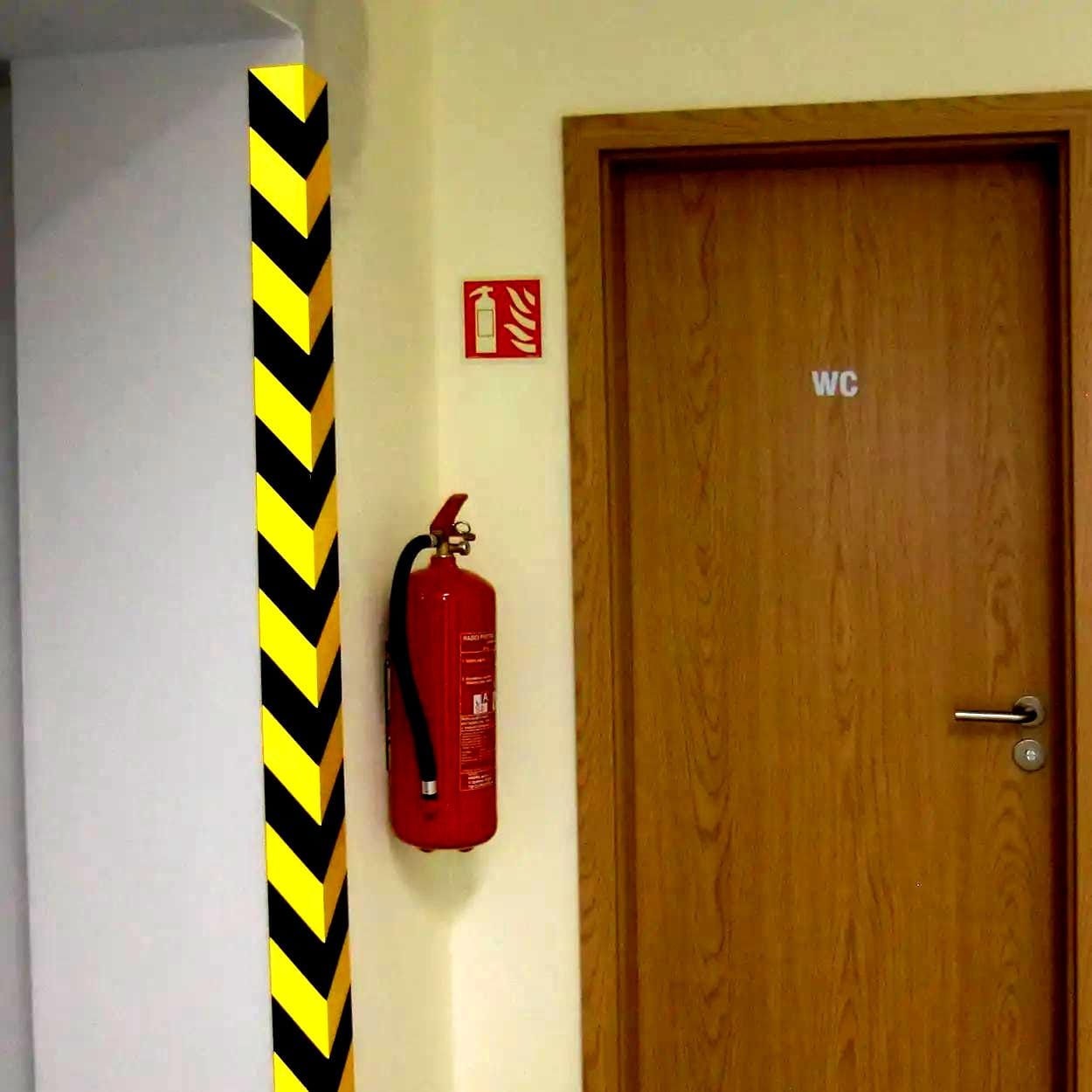Widok na drzwi do toalety oznaczone „WC”, obok stojąca gaśnica oraz słup ze wzorem ostrzegawczym w kolorze czarno-żółtym. Elementy te podkreślają bezpieczeństwo pracy i środki awaryjne w budynku.