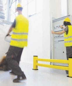 Pracownicy w żółtych kamizelkach odblaskowych i kaskach wykonują prace w magazynie, transportując materiały w pobliżu bariery ochronnej. Scena ilustruje zasady bezpieczeństwa w miejscu pracy oraz zastosowanie środków awaryjnych w celu ochrony przed wypadkami.