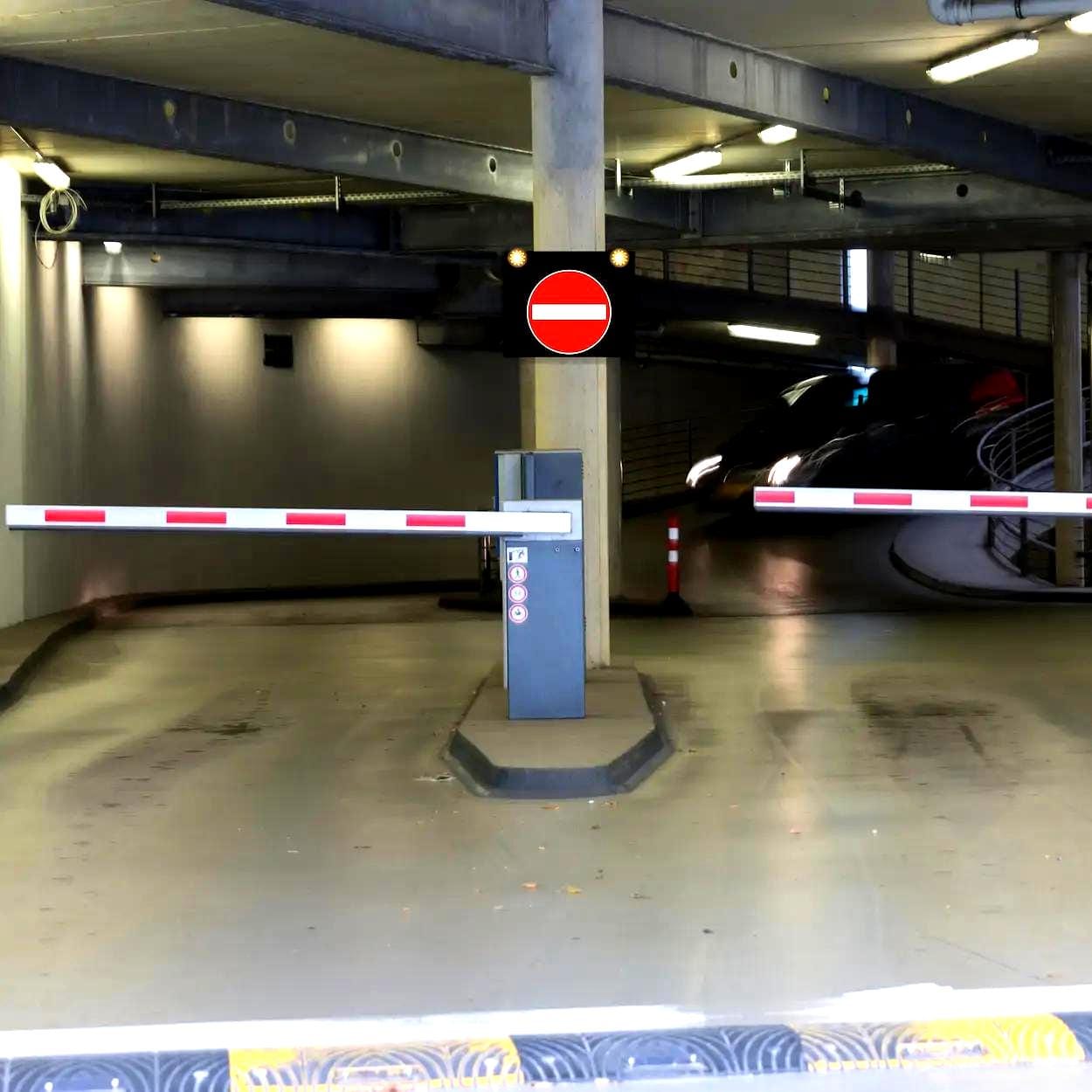 Widok na wjazd do parkingu z zaporą i znakiem zakazu wjazdu. Na drodze widoczne są oznaczenia, a w tle widać wyjeżdżający samochód. Obszar ten ilustruje zasady bezpieczeństwa pracy i zarządzania ruchem w przestrzeni publicznej.