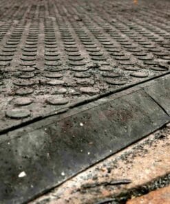 Zbliżenie na brudną nawierzchnię z wypukłymi okrągłymi elementami, która jest częściowo pokryta zanieczyszczeniami. Widoczna krawędź z materiału gumowego, co może sugerować zastosowanie w kontekście bezpieczeństwa pracy lub ochrony środowiska.