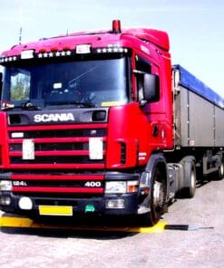 Czerwony samochód ciężarowy Scania z przyczepą na drodze, wyposażony w dodatkowe oświetlenie. W tle widoczna zieleń, co sugeruje dbałość o ochorona środowiska. Przykład nowoczesnego transportu oraz znaczenia bezpieczeństwa pracy w logistyce.