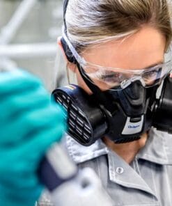 Kobieta w masce ochronnej i goglach laboratoryjnych, wykonująca eksperymenty w laboratorium, nosi rękawice ochronne. Obraz ilustruje ważność bezpieczeństwa pracy i stosowanie środków awaryjnych w kontekście ochrony środowiska.