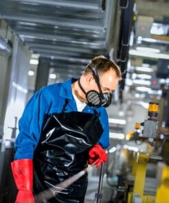 Mężczyzna w niebieskiej koszuli roboczej i czarnej odzieży ochronnej pracuje w fabryce, nosząc maskę ochronną i rękawice. Skupia się na swoim zadaniu, co podkreśla znaczenie bezpieczeństwa pracy i ochrony środowiska w przemyśle.