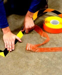 Mężczyzna umieszcza taśmę ostrzegawczą w czarnych i żółtych kolorach na podłodze. Obok leży rolka pomarańczowej taśmy. Aktywności związane z bezpieczeństwem pracy w celu ochrony środowiska i zapobiegania wypadkom.