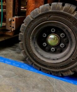 Widok na oponę wózka widłowego stojącego na niebieskiej matce ochronnej, otoczonego drewnianymi belkami w tle. Opona ma wyraźne żłobienia, co zapewnia bezpieczeństwo pracy w magazynie i chroni powierzchnię przed uszkodzeniami.