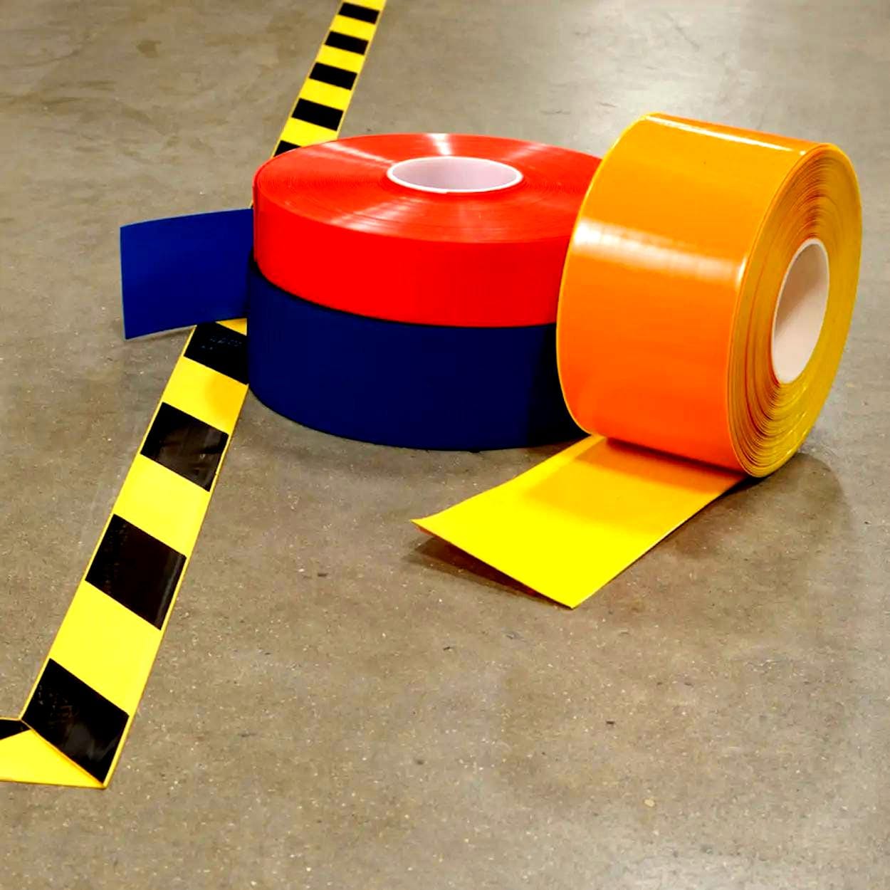 Tasma w różnych kolorach: czerwona, niebieska, żółta i pomarańczowa, leży na betonowej podłodze obok czarno-żółtej linii. Używane w środki awaryjne do oznaczania i bezpieczeństwa pracy w magazynach i zakładach przemysłowych.