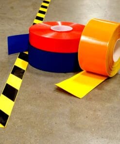 Zestaw kolorowych taśm ostrzegawczych na betonowej podłodze, w tym czerwonej, niebieskiej, żółtej i czarnej w paski, idealny do oznakowania obszarów roboczych oraz poprawy bezpieczeństwa pracy w środowisku produkcyjnym.