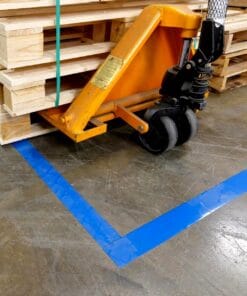 Złota wózek paletowy z dużymi kołami, umieszczony obok stosu drewnianych palet, na wyłożonym niebieskim pasem podłogi w magazynie. Ujęcie ukazuje szczegóły narzędzia i oznakowanie podłogi, istotne dla bezpieczeństwa pracy i organizacji przestrzeni.