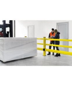 Pracownicy w magazynie omawiają dokumenty przy żółtych barierkach ochronnych, podczas gdy w tle widać wózek widłowy przewożący zapakowany towar. Scena ukazuje bezpieczeństwo pracy i organizację w środowisku magazynowym.