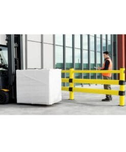 Mężczyzna w pomarańczowej kamizelce bezpieczeństwa przy kontrolowaniu palety z towarem za pomocą wózka widłowego, obok żółta barierka ochronna na terenie zakładu przemysłowego. Zdjęcie ilustruje zasady bezpieczeństwa pracy i ochrony środowiska w logistyce.