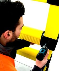 Mężczyzna w pomarańczowej kamizelce roboczej montuje elementy metalowe za pomocą wkrętarki. W tle widoczne są żółte konstrukcje, co podkreśla znaczenie bezpieczeństwa pracy i stosowanie odpowiednich środków awaryjnych podczas prac konstrukcyjnych.