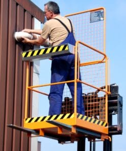 Mężczyzna w niebieskim kombinezonie roboczym, stojący na podnośniku, montuje element na ścianie budynku. W tle jasne niebo. Prace na wysokości wymagają szczególnej ostrożności, co podkreśla znaczenie bezpieczeństwa pracy i ochrony środowiska.