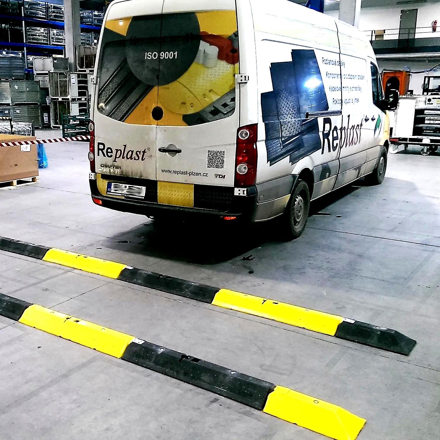 Widzimy samochód dostawczy z logo Replast w magazynie. Na podłodze znajdują się progi z żółto-czarnymi oznaczeniami, które zwiększają bezpieczeństwo pracy w terenie. W tle widać regały z towarami, co wskazuje na aktywne środowisko przemysłowe.