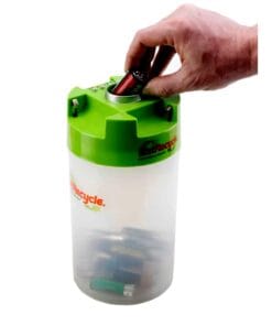 Pojemnik do recyklingu baterii z zielonym wiekiem, do którego wkładana jest bateria. W środku widoczne są zużyte baterie, co podkreśla znaczenie recyklingu w ochronie środowiska.
