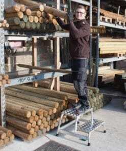 Mężczyzna stojący na drabinie, trzymający drewnianą belkę w drewnianym magazynie, otoczony przez stosy bali na półkach. Scena ilustruje znaczenie bezpieczeństwa pracy oraz ergonomicznych metod w branży budowlanej, wspierających ekologię i ochronę środowiska.