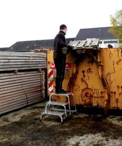 Mężczyzna staje na drabince, aby umieścić deski w kontenerze na odpady. W tle widoczne palety i oznaczenia dotyczące bezpieczeństwa. Scena związana z gospodarką odpadami i recyklingiem.