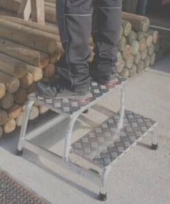 Osoba stająca na metalowej drabinie, wykonanej z materiału antypoślizgowego, obok stosu drewnianych bali. Drabina jest stabilna, co zapewnia bezpieczeństwo pracy w trakcie wykonywania zadań wysokościowych. Przykład użycia sprzętu w praktyce związany z ekologią i ochroną środowiska.