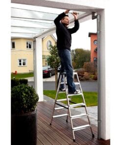 Mężczyzna w czarnym swetrze stoi na drabinie, naprawiając konstrukcję z przeszklonym dachem. W tle widać zadbane ogrody i nowoczesne budynki, co podkreśla dbałość o ekologię i bezpieczeństwo pracy w obszarze budownictwa.