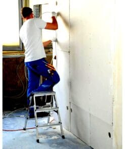 Mężczyzna pracuje na drabince, montując ścianki kartonowo-gipsowe w pomieszczeniu budowlanym, dbając o bezpieczeństwo pracy w trakcie remontu. W tle widać okno z naturalnym światłem oraz nieuporządkowaną podłogę z narzędziami i przewodami.