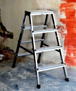 Szeroka drabina aluminiowa ustawiona w pomieszczeniu budowlanym obok czerwonej, świeżo malowanej ściany. W tle widoczne prace remontowe oraz kable elektryczne, co podkreśla kontekst bezpieczeństwa pracy i użycia odpowiednich środków awaryjnych w trakcie renowacji.