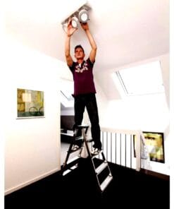 Mężczyzna stoi na drabinie, montując oświetlenie sufitowe w jasnym wnętrzu mieszkania. Jego działania ilustrują zagadnienia związane z bezpieczeństwem pracy oraz użyciem odpowiednich środków awaryjnych w trakcie prac remontowych.