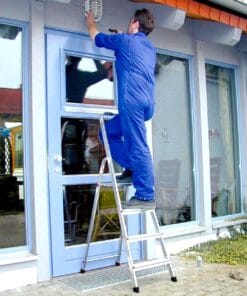 Mężczyzna w ogrodniczkach montuje lampę na zewnątrz budynku, stojąc na drabince przy drzwiach. Zwrócony jest w kierunku górnej części drzwi, co podkreśla znaczenie bezpieczeństwa pracy podczas prac na wysokości i dbałość o estetykę otoczenia.