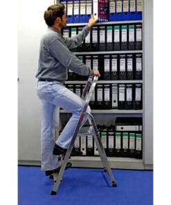 Mężczyzna stojący na drabinie, sięgający po segregator z górnej półki w biurze. W tle widoczne uporządkowane segregatory w różnych kolorach, co podkreśla znaczenie organizacji dokumentów w kontekście bezpieczeństwa pracy i efektywności.