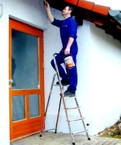 Mężczyzna w niebieskim kombinezonie maluje sufit pod dachówką domu, stojąc na drabinie. W jednej ręce trzyma pędzel, w drugiej puszkę farby, co ilustruje prace remontowe i dbałość o środowisko w kontekście bezpieczeństwa pracy.