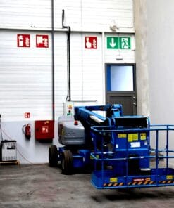 Zimny, przestronny magazyn z niebieską podnośnikiem oraz znakami bezpieczeństwa awaryjnego. Widoczne są sprzęty gaśnicze i infrastruktura ochrony środowiska. Malowana podłoga z wyraźnym podziałem przestrzeni roboczej, co sprzyja bezpieczeństwu pracy.